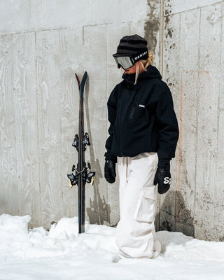 Person in winter clothing holding skis against a concrete wall with snow on the ground